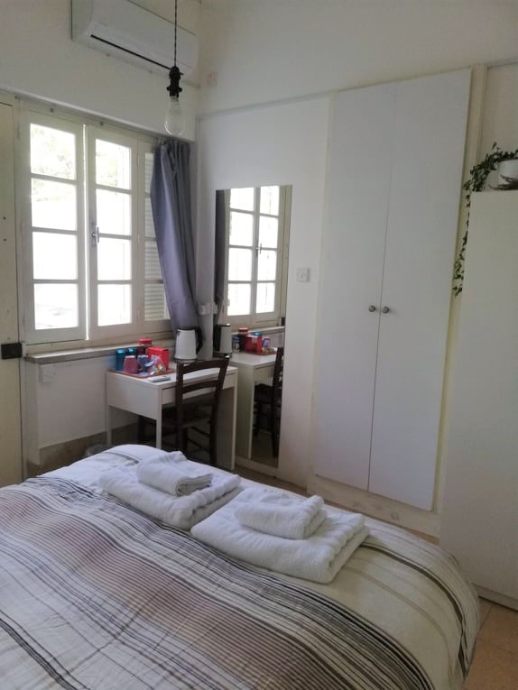 Bedroom view from the bed, a small talbe with cups and kettle, air condition on the wall and big rectangle mirror, the white door to the bathroom, dresser with green plant on the top
